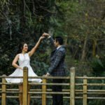 Bride in a short white dress and groom in a checkered suit dancing happily on a wooden bridge surrounded by lush green trees
