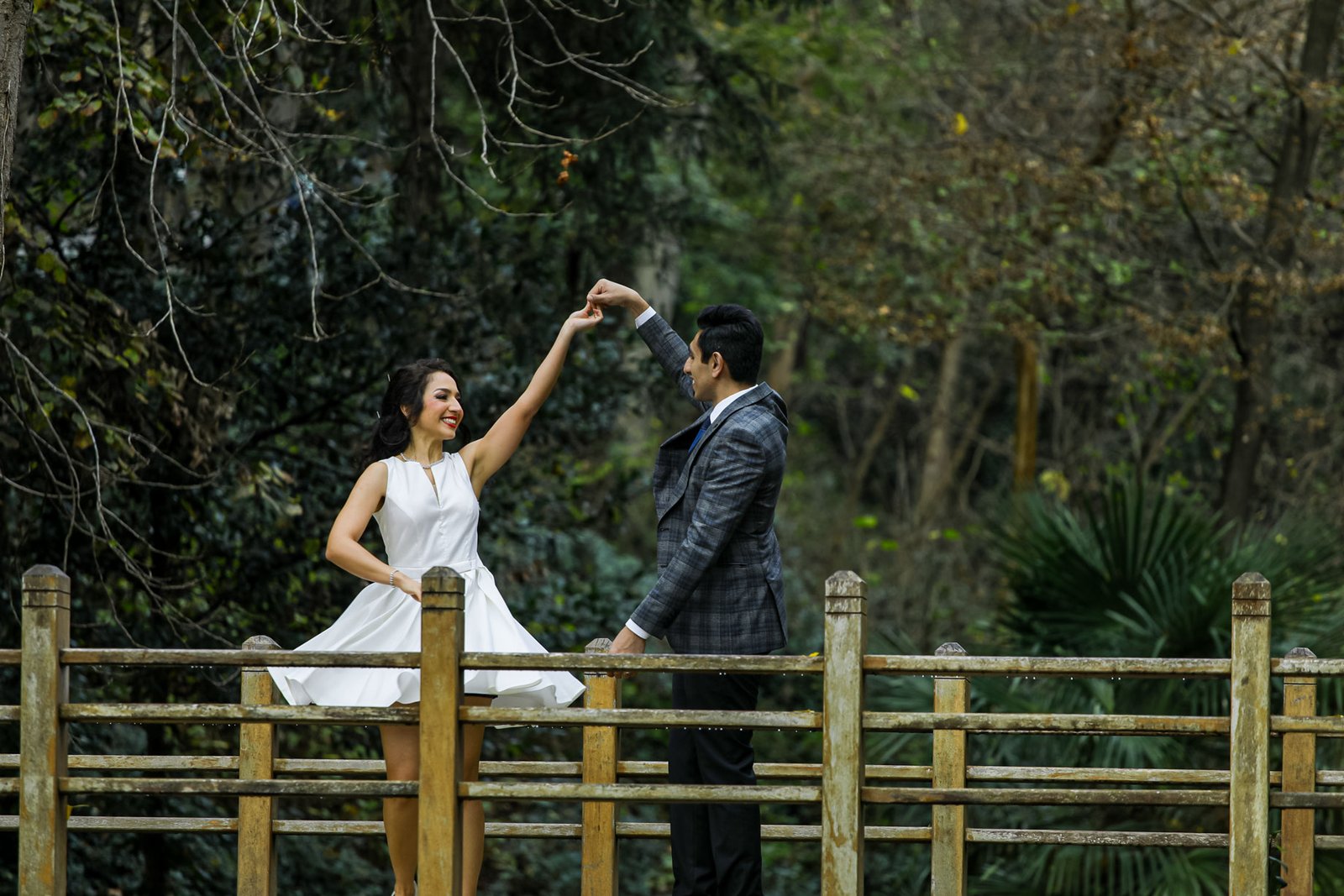 Bride in a short white dress and groom in a checkered suit dancing happily on a wooden bridge surrounded by lush green trees