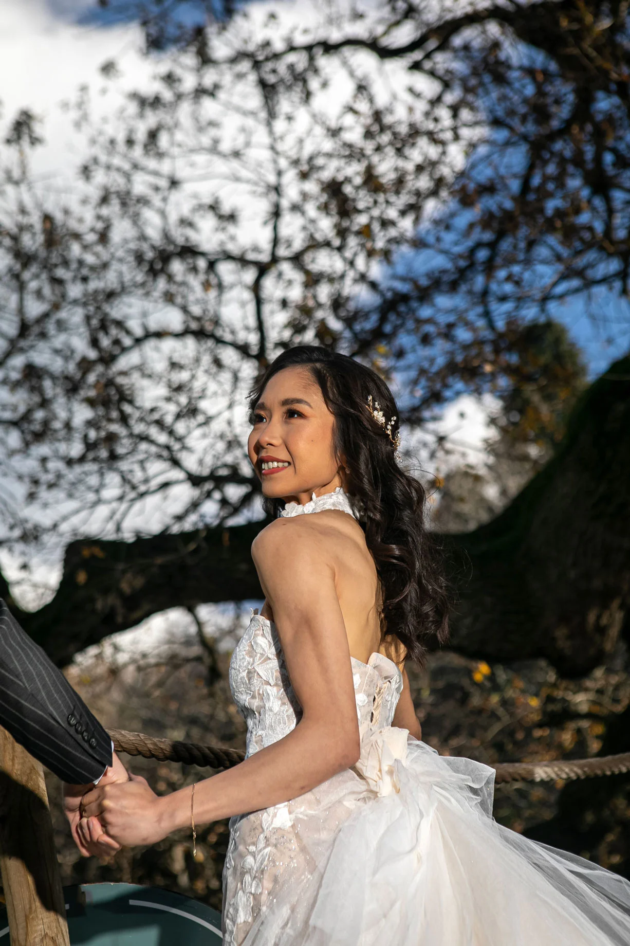 Smiling Asian bride in a lace wedding dress holding hands with groom during outdoor wedding photoshoot